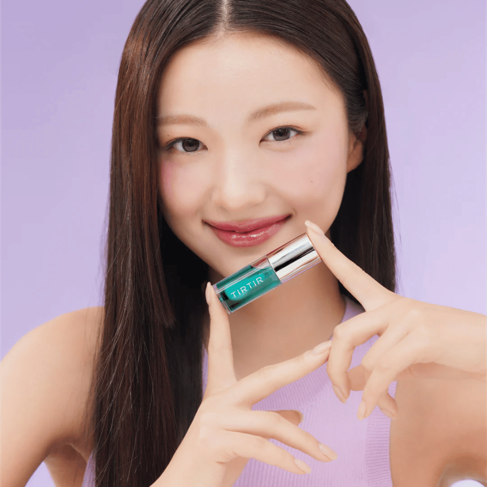 Nainen, jolla on pitkät, suorat hiukset, pitää kädessään TIRTIR My Glow Lip Oil ja esittelee kiiltäviä, vaaleanpunaisia huuliaan violettia taustaa vasten.