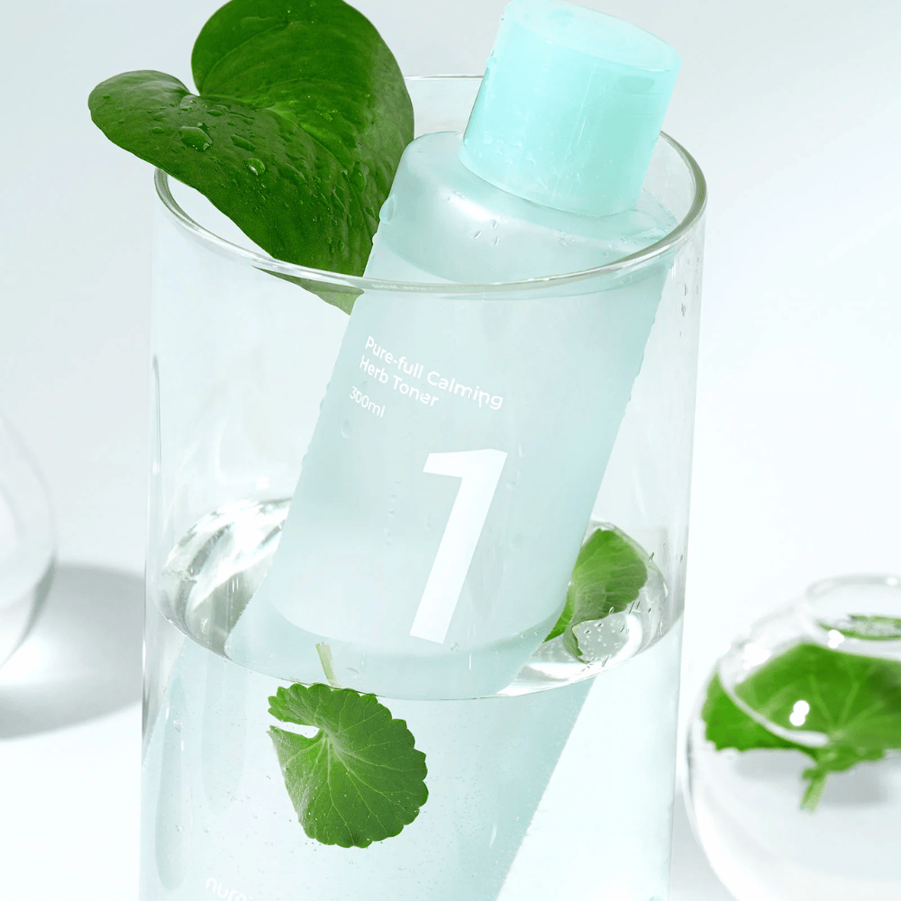 Pullo, jossa on No.1 Pure-Full Calming Herb Toner, sijoitettuna lasiin, jossa on vettä ja vihreitä lehtiä. Pullossa on vaaleansininen korkki ja etiketti, jossa lukee "Pure-full Calming Herb Toner" sekä "300ml". Tuote näyttää raikkaalta veden ja kasvillisuuden taustalla, mikä antaa rauhoittavan ja luonnollisen tunteen.