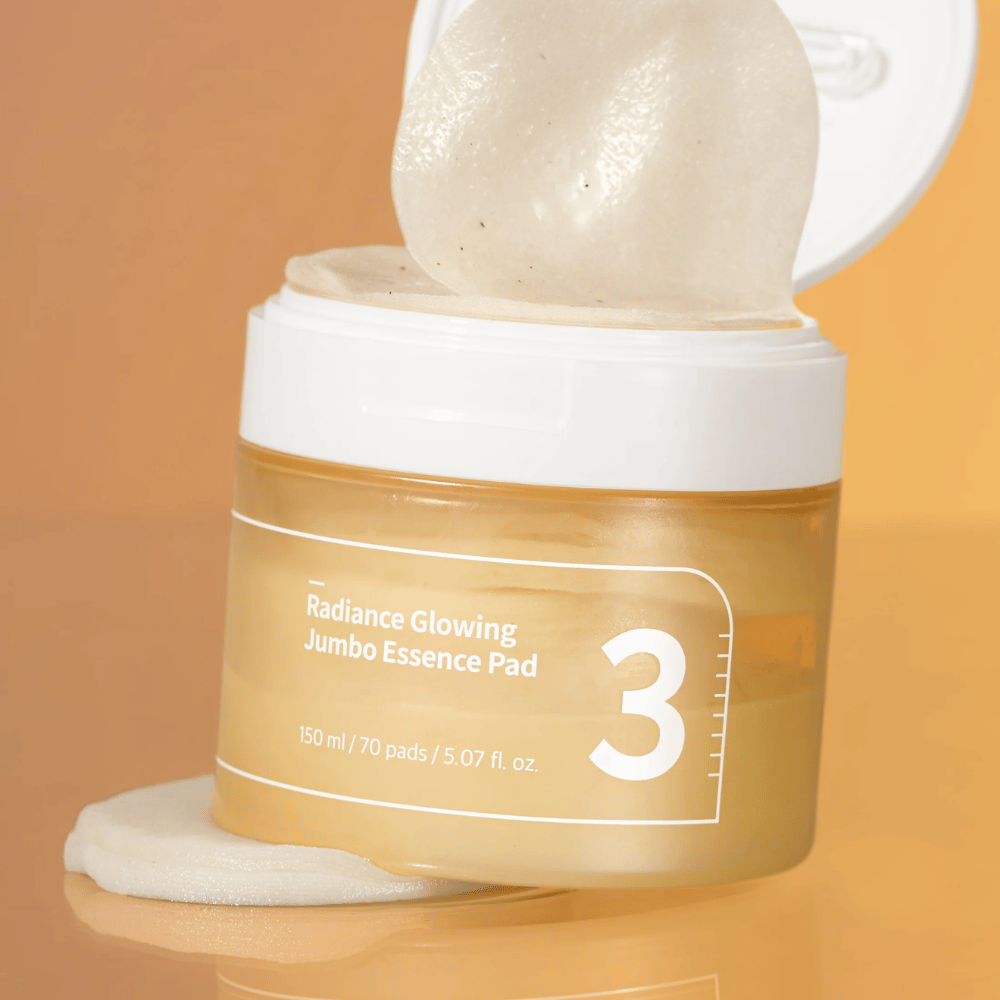 Avoin purkki, jossa on numbuzin No. 3 Radiance Glowing Jumbo Essence Pad, yksi tyyny nojaa reunaa vasten, taustalla oranssi sävy.