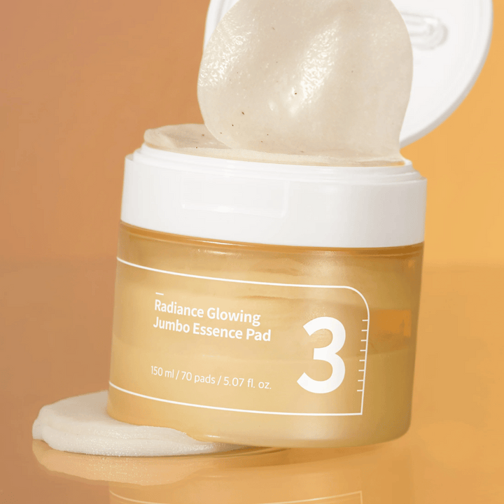 Öppen burk med numbuzin No. 3 Radiance Glowing Jumbo Essence Pad, en pad syns lutad mot kanten, bakgrund i orange ton.