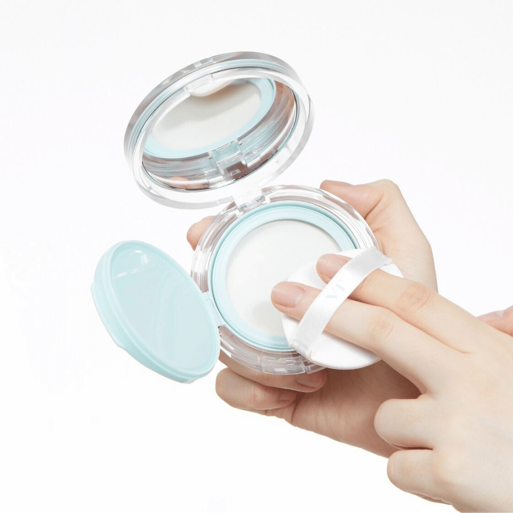 Käsi pitelee avointa purkkia VT PDRN Essence Glow Sun Pact:llä. Puffi on matkalla levittämään puuteria, jossa on valkoinen pinta ja sininen reuna purkin ympärillä.