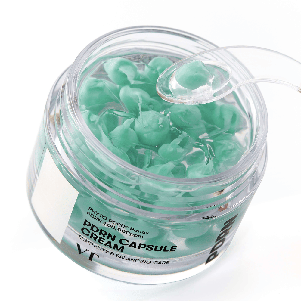 Lähikuva avoimesta VT PDRN Capsule Cream -purkista. Sisältö koostuu pienistä vihreistä kapseleista läpinäkyvässä geelissä, ja mukana on spaatteli levitystä varten. Purkin tekstissä lukee "Elasticity & Balancing Care".
