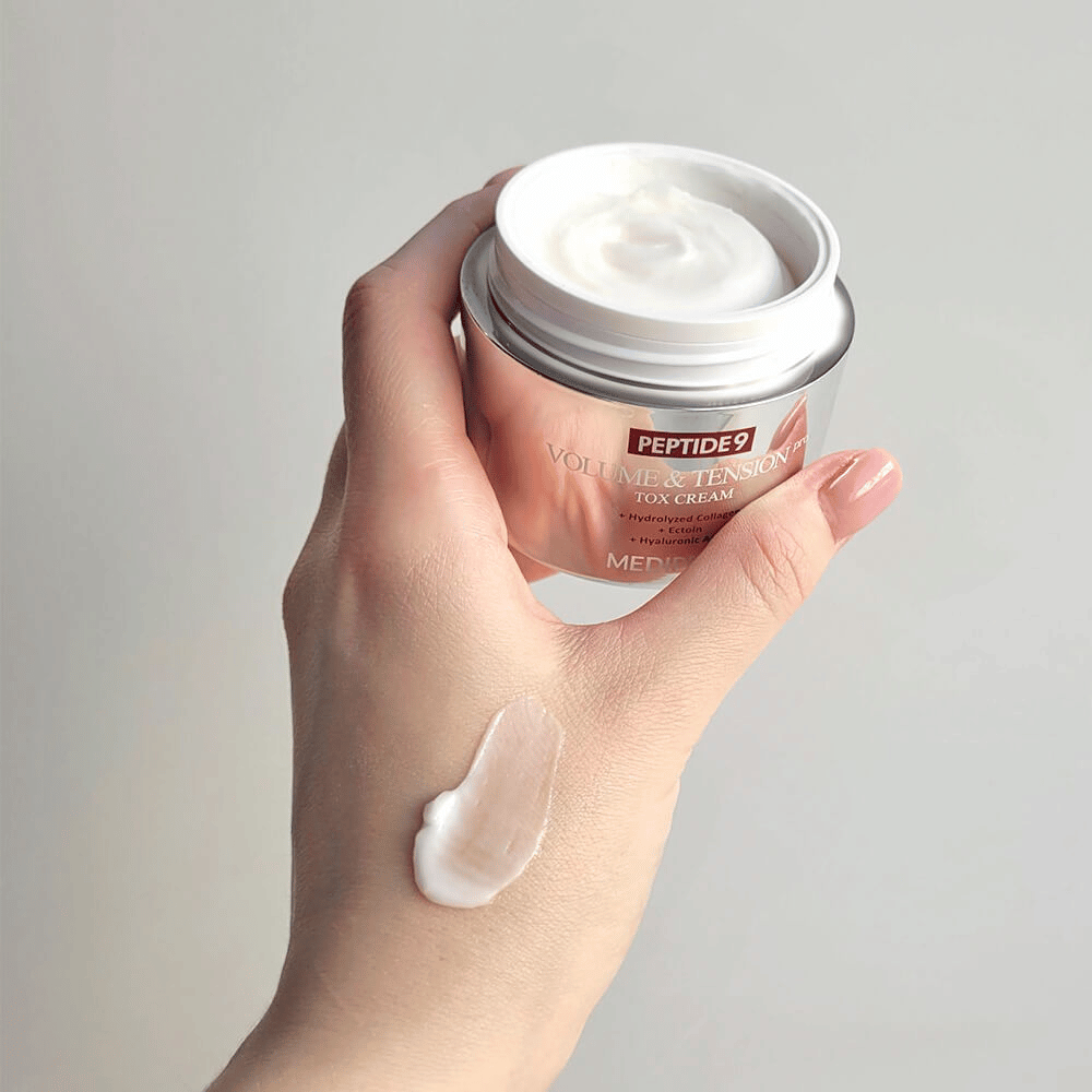 Käsi pitää MEDI-PEEL Peptide 9 Volume & Tension Tox Cream -voidetta, jossa on voiteen nokare kämmenselällä. Silkkinen koostumus kosteuttaa ja antaa iholle kiinteyttä ja kimmoisuutta.