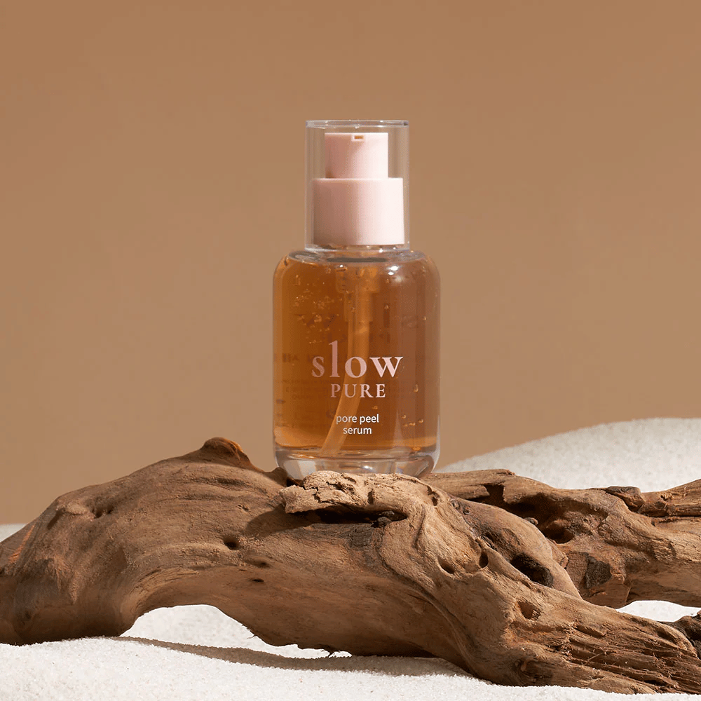 Slow Pure Pore Peel Serum sijoitettuna hiekalle puuyksityiskohta taustalla, symboli luonnolliselle ihonhoidolle.