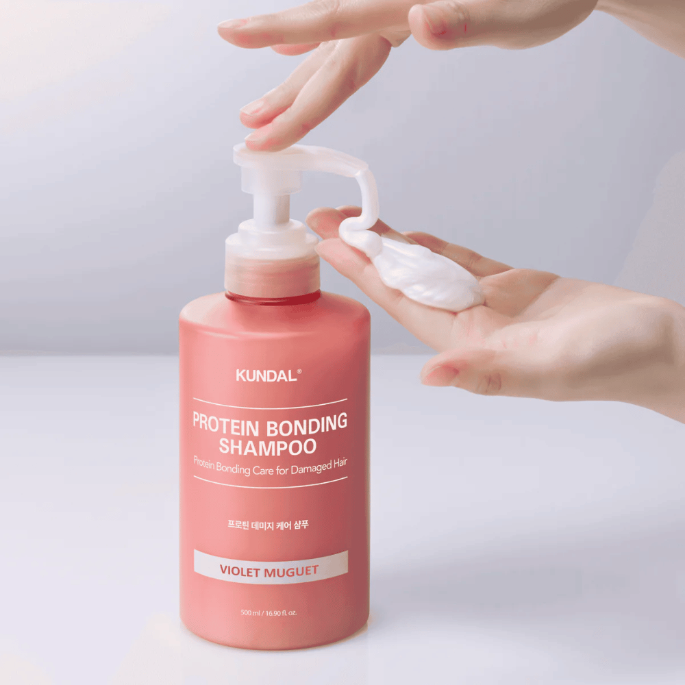 Käsi pumppaa ulos KUNDAL Protein Bonding Shampoo Violet Muguet kermaisessa koostumuksessa, korjaava shampoo vaurioituneille hiuksille.