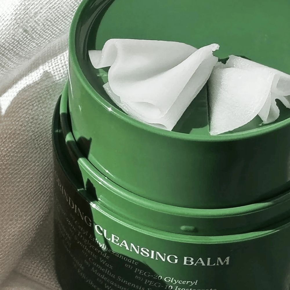 Kuva näyttää vihreän purkin, jossa on "Cleansing Balm", joka on avattu paljastamaan mukana tulevan lastan ja pehmeän sisällön. Kermainen koostumus heijastaa valoa, mikä antaa ylellisyyden ja tehokkuuden tunteen puhdistuksessa.