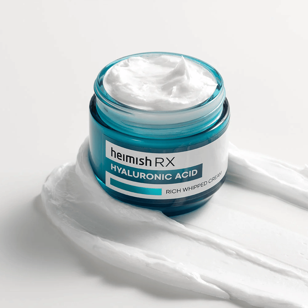 Heimish RX Hyaluronic Acid Rich Whipped Cream sinisessä lasipurkissa, jossa on rikas ja kuohkea koostumus, levitettynä valkoiselle taustalle. Intenssiivisesti kosteuttava kasvovoide, joka sisältää hyaluronihappoa syvään kosteutukseen ja pehmeälle iholle.
