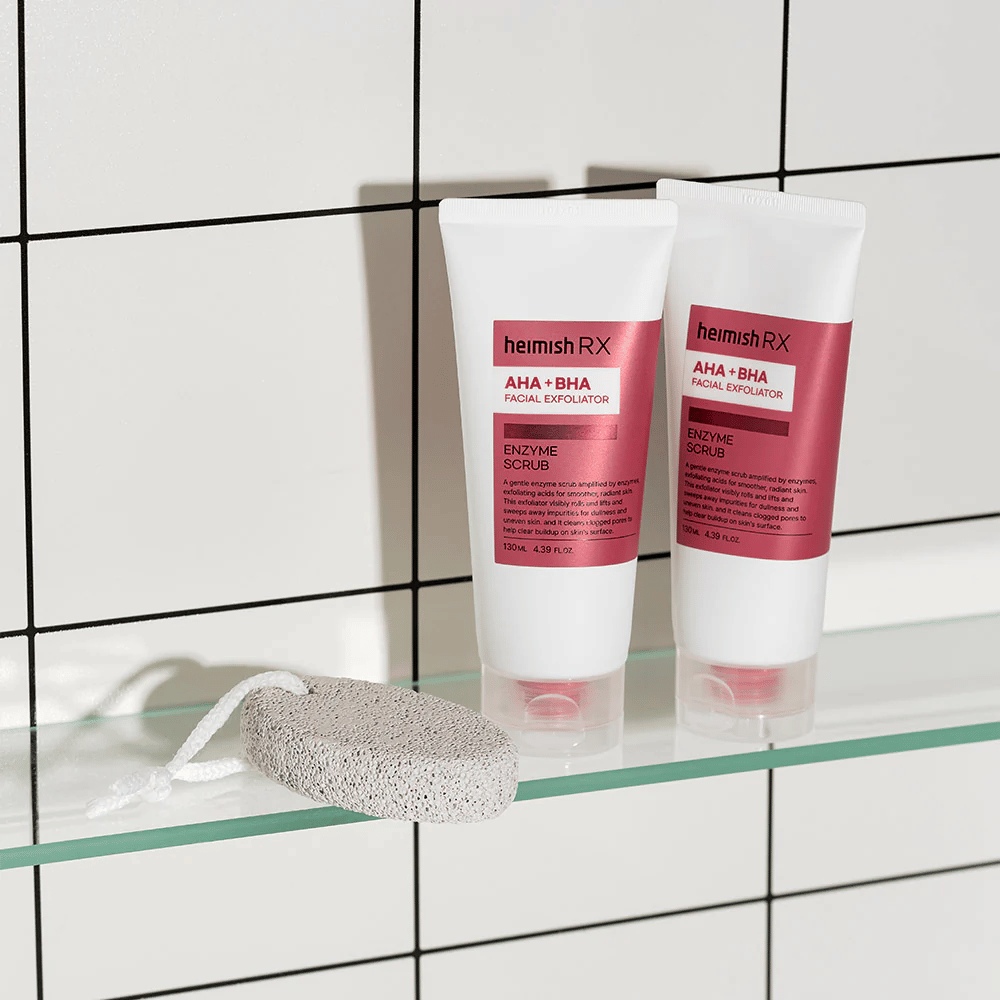Kaksi tuubia Heimish RX AHA + BHA Facial Exfoliator -tuotetta asetettuna lasihyllylle kylpyhuoneessa, jossa on valkoinen kaakeliseinä. Tuotteiden vieressä on hohkakivi, jossa on valkoinen naru. Tyylikäs ja minimalistinen ihonhoitokuva.