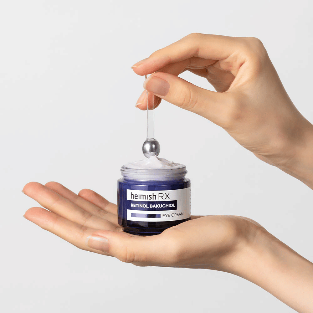Heimish RX Retinol Bakuchiol Eye Cream tyylikkäässä sinisessä purkissa, nostetaan metallisella applikaattorilla. Silmänympärysvoide, jossa on retinolia ja bakuchiolia hienojen juonteiden vähentämiseksi ja kiinteämmän ihon saamiseksi.