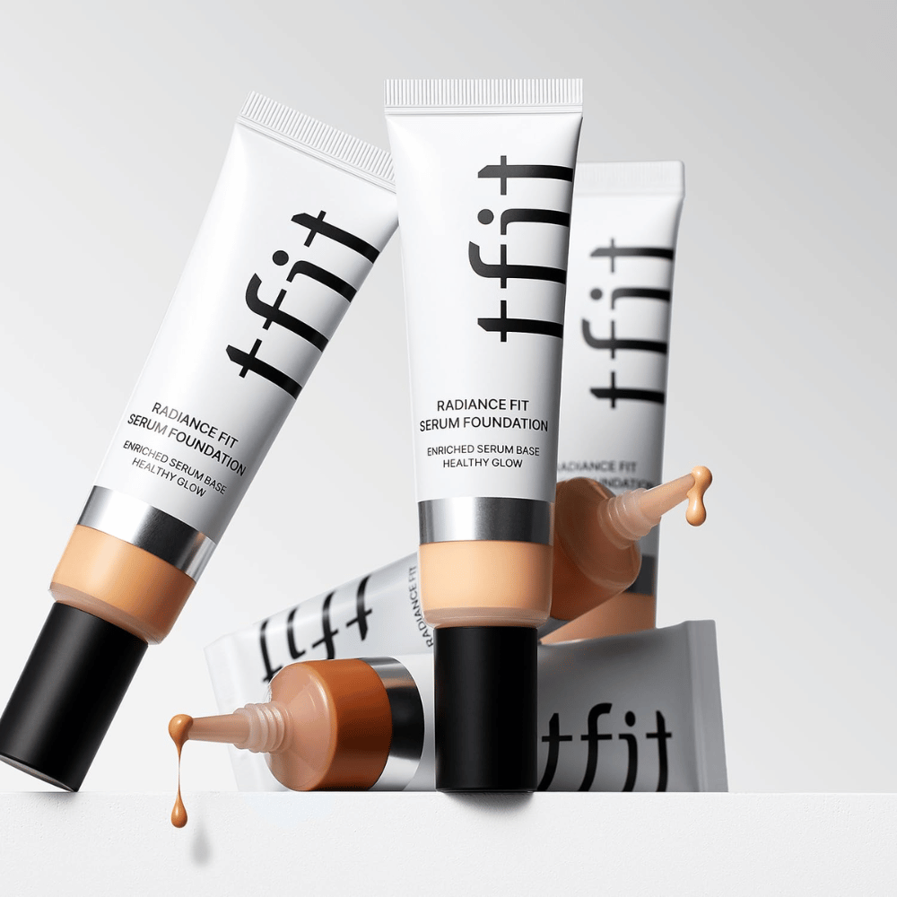 Useita TFIT Radiance Fit Serum Foundation -tuubeja eri sävyissä, joista yhdestä foundation tippuu, vaaleanharmaata taustaa vasten.