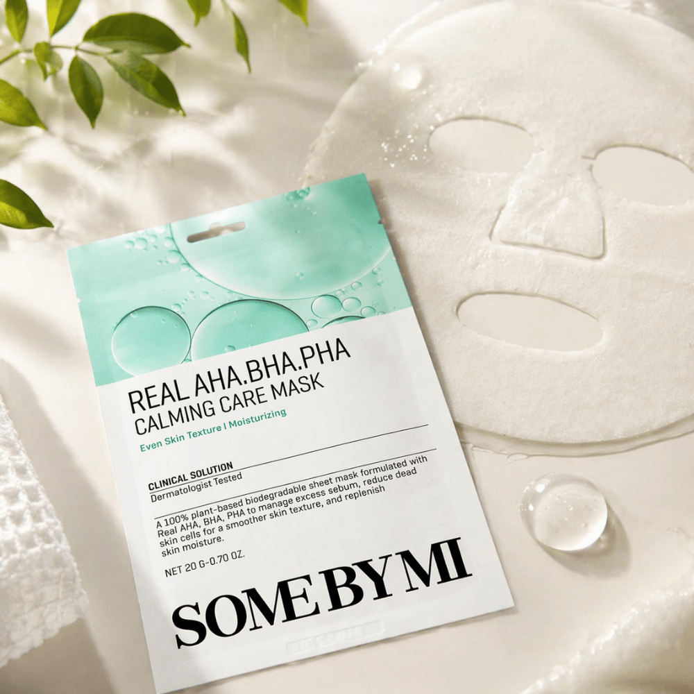 SOME BY MI Real AHA.BHA.PHA Calming Care Mask esitetään yhdessä avattuna olevan kangasnaamion kanssa. Tuotteen ympärillä näkyy vesipisaroita ja vihreitä lehtiä, jotka vahvistavat kosteutuksen, rauhoittavan hoidon ja hellävaraisen kuorinnan tuntua. Kuva korostaa naamion keskittymistä tasaisempaan ihon tekstuuriin ja parantuneeseen kosteustasapainoon.