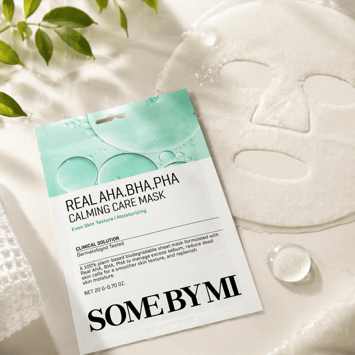 SOME BY MI Real AHA.BHA.PHA Calming Care Mask esitetään yhdessä avattuna olevan kangasnaamion kanssa. Tuotteen ympärillä näkyy vesipisaroita ja vihreitä lehtiä, jotka vahvistavat kosteutuksen, rauhoittavan hoidon ja hellävaraisen kuorinnan tuntua. Kuva korostaa naamion keskittymistä tasaisempaan ihon tekstuuriin ja parantuneeseen kosteustasapainoon.
