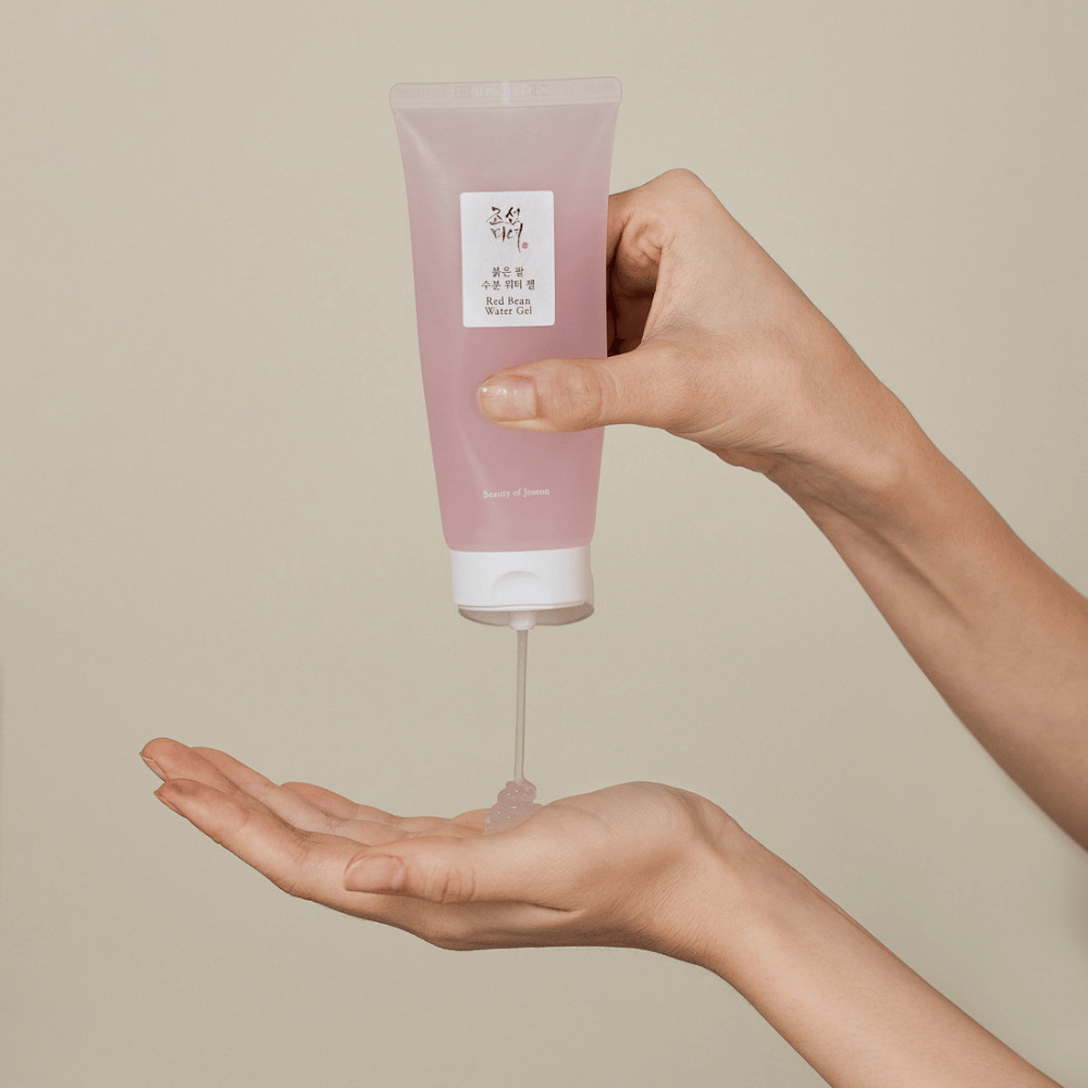 Kuva näyttää henkilön, joka pitää kädessään putkiloa "Red Bean Water Gel" Beauty of Joseon ja puristaa läpinäkyvää geeliä käteensä. Tuote näyttää olevan tarkoitettu ihonhoitoon ja sillä on houkutteleva, pehmeän vaaleanpunainen pakkaus.