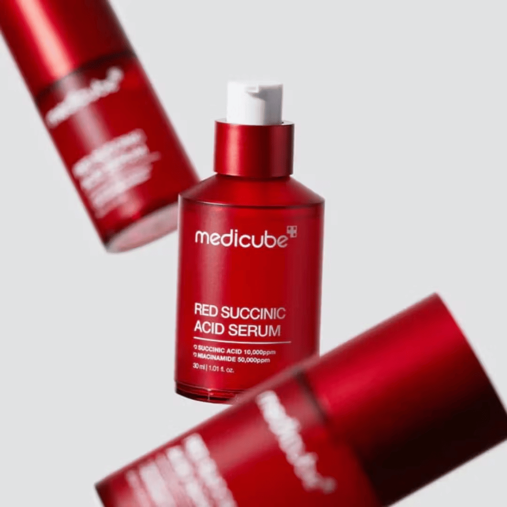 Medicube Red Succinic Acid Serum punaisessa pumppupullossa, ympäröitynä useilla identtisillä pulloilla vaaleaa taustaa vasten. Tuote sisältää sukiniinihappoa ja niasiiniamidia aknen ja pigmenttiläiskien hoitoon.