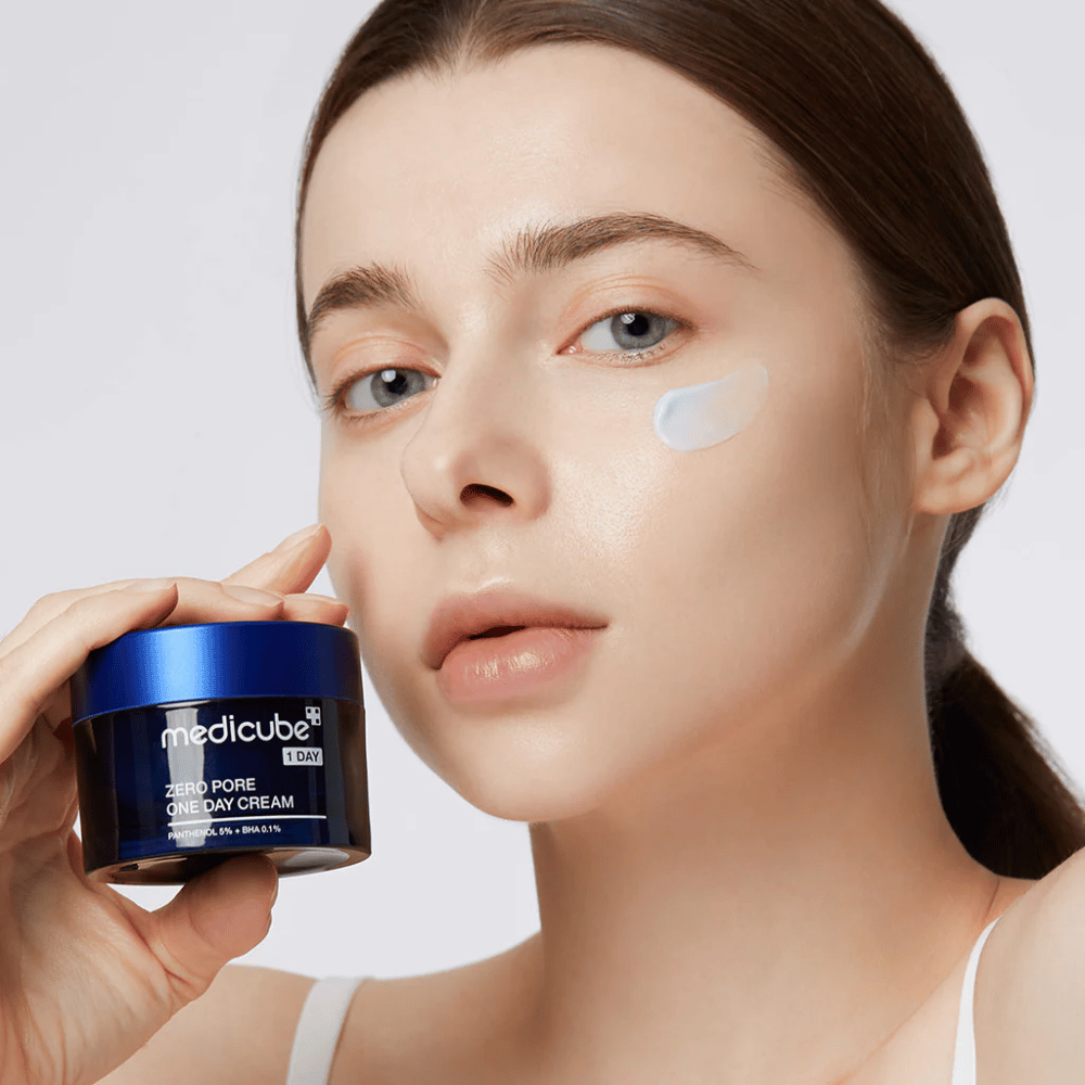 Nainen luonnollisella ilmeellä pitää Medicube Zero Pore One Day Cream. Hänen poskelleen on levitetty pieni määrä voidetta. Tuote on suunniteltu parantamaan ihon rakennetta ja minimoimaan huokoskokoa kevyellä ja kosteuttavalla koostumuksella.
