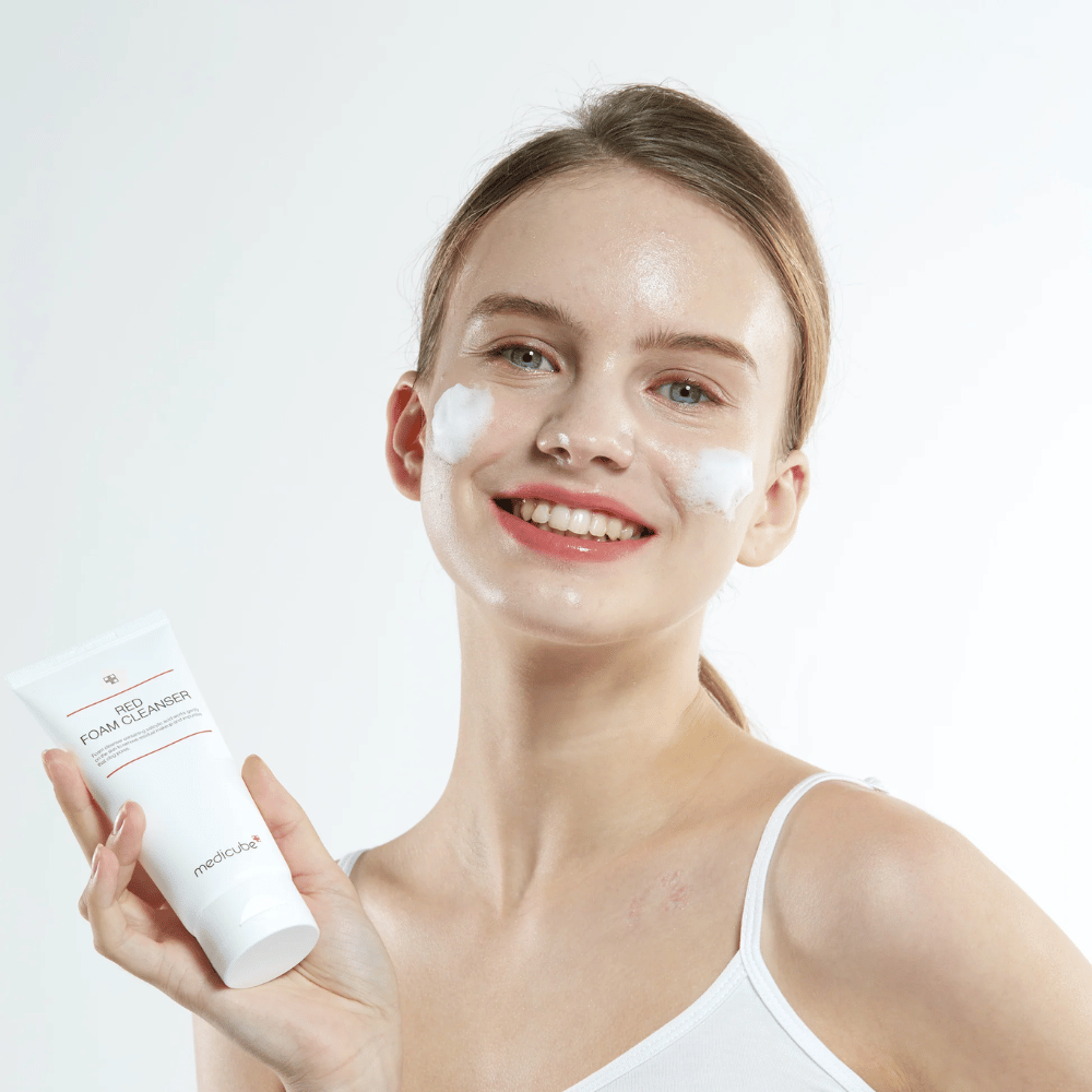 Nuori nainen, jolla on puhdistusvaahtoa kasvoillaan, pitää kädessään Medicube Red Foam Cleanser -tuubia ja hymyilee kameralle. Hänellä on yllään valkoinen toppi ja tausta on valoisa ja yksinkertainen.