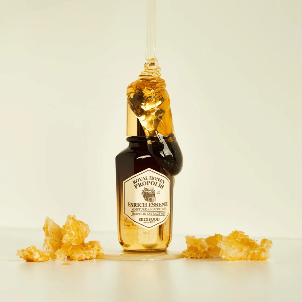 Kuva lasipullosta, jossa on Royal Honey Propolis Enrich Essence Skinfood. Suuri kultaisen hunajan pisara valuu pullon kultaisesta korkista. Pullon ympärillä on pieniä hunajakennon palasia hajallaan vaalealla pinnalla. Essenssiä markkinoidaan keskittyen kosteuteen ja ravintoon, 63% propolisuutetta.
