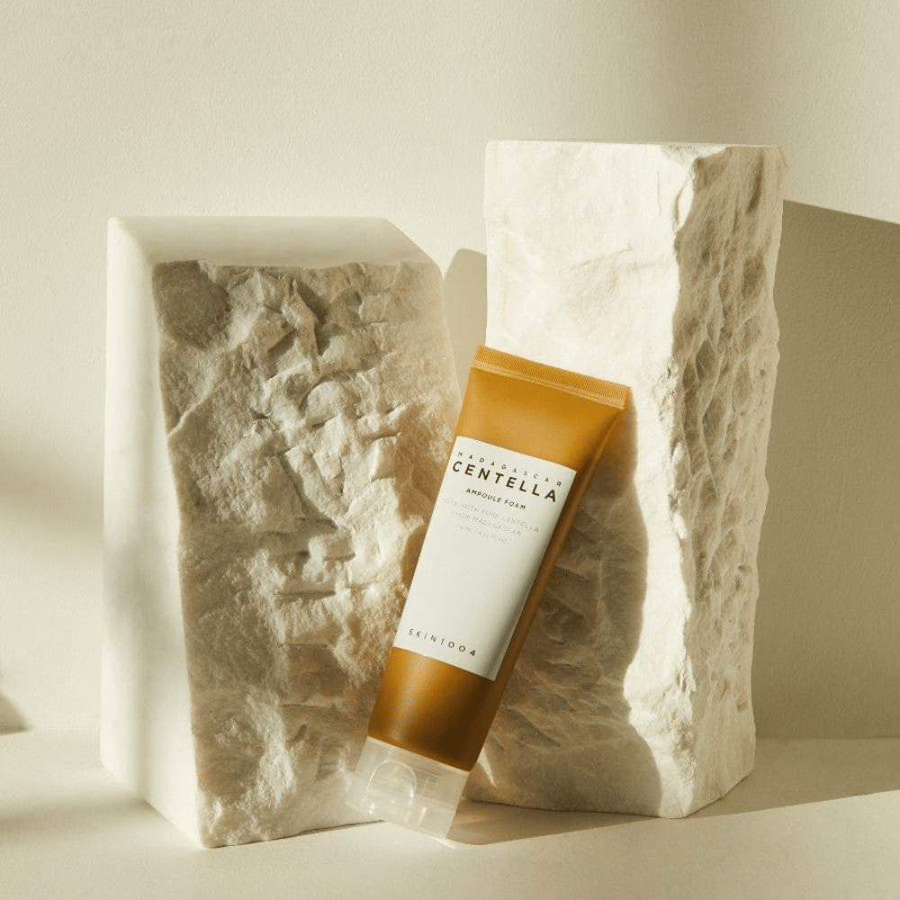 SKIN1004 Madagascar Centella Ampoule Foam seisoo vaaleita kiviblokkeja vasten luonnonvalossa, ruskeavalkoisella tuubimuotoilullaan ja elegantilla minimalismillaan.