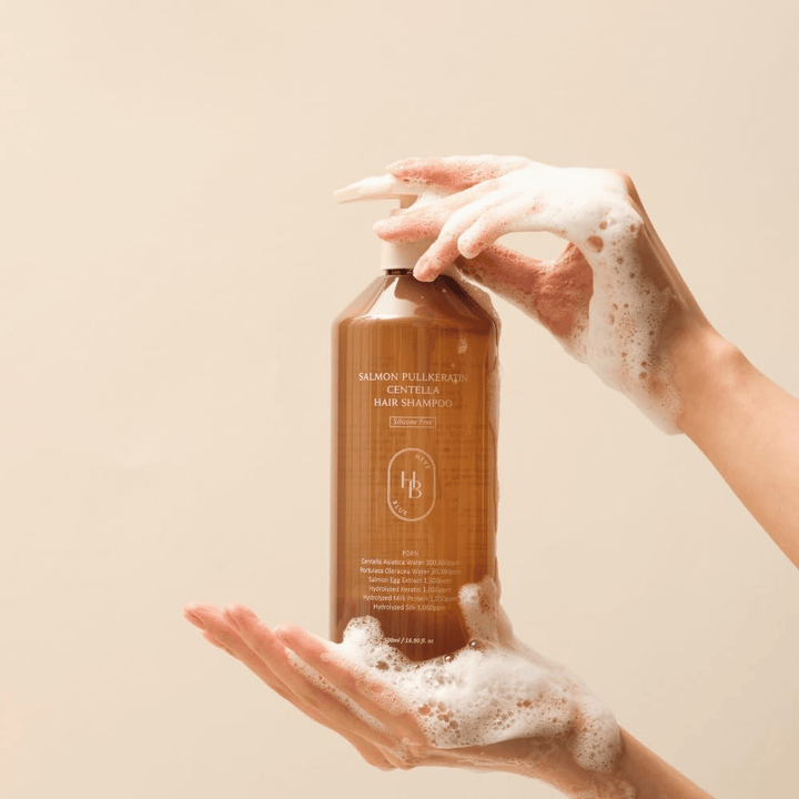 Heveblue Salmon Pullkeratin Centella Hair Shampoo ruskeassa pullossa vaahdon ympäröimänä. Tuotetta pidetään kahden käden välissä, ja se korostaa puhdistavaa ja hoitavaa hiustenhoitokokemusta runsaalla vaahdolla.