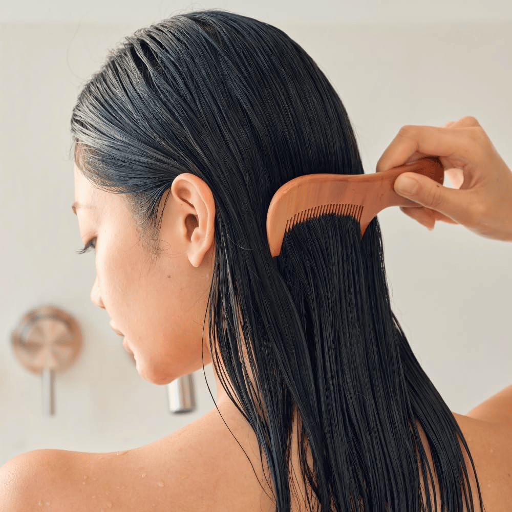 Henkilö kampaa pitkiä, märkiä hiuksia leveähampaisella kammalla levitettyään Heveblue Salmon Pullkeratin Centella Hair Treatment. Havainnollistaa selvittävää ja pehmentävää vaikutusta sileämmän lopputuloksen saavuttamiseksi.