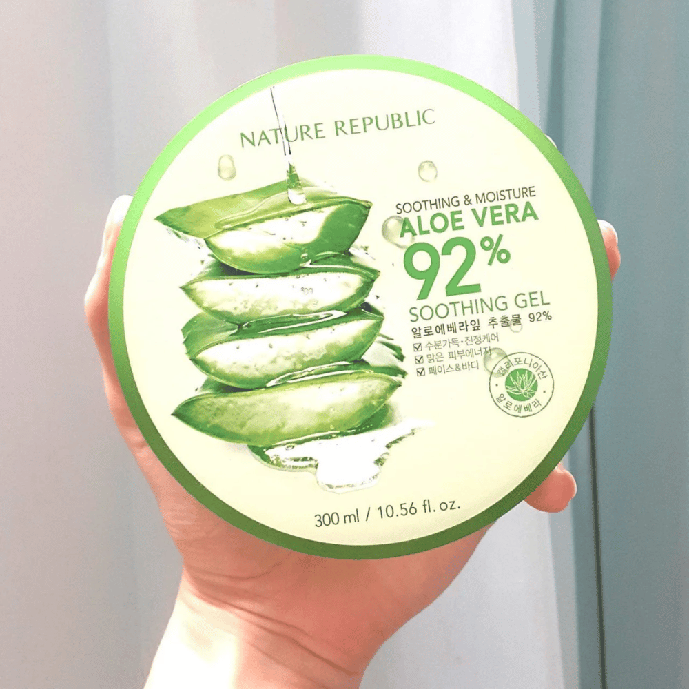 Käsi pitää ylhäällä pyöreää purkkia, jossa on Nature Republic Aloe Vera 92% Soothing Gel. Purkki on vihreä ja siinä on kuva aloe verasta sekä teksti, joka kuvaa sen rauhoittavia ja kosteuttavia ominaisuuksia. Sisältö on 300 ml.