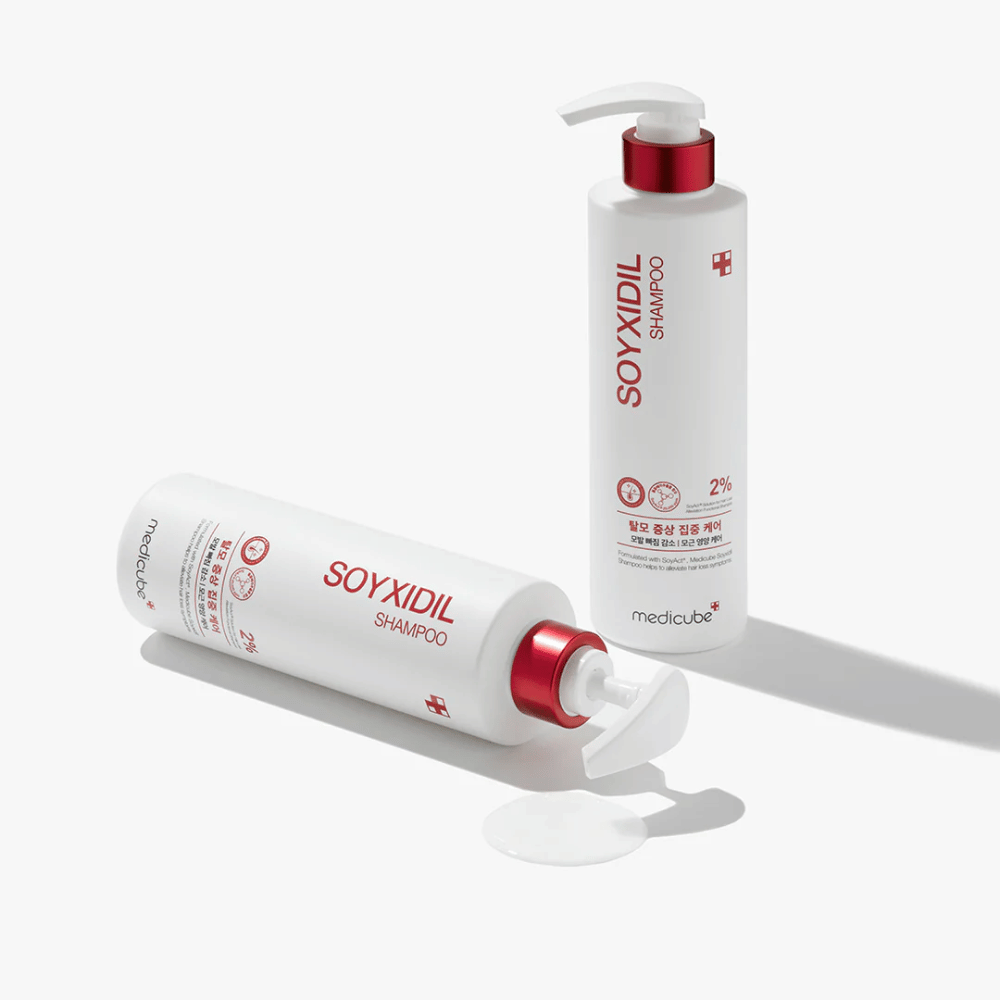 Kaksi pulloa Medicube Soyxidil Shampoo valkoisessa ja punaisessa designissa, yksi pystyasennossa ja toinen makuuasennossa, shampooa läikkyneenä vieressä.