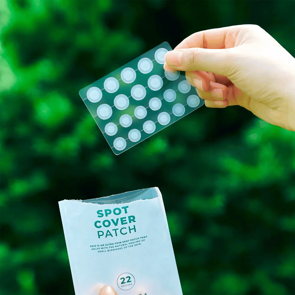 Käsi pitelee Spot Cover Patches -arkkia, jotka ovat läpinäkyviä ja pyöristettyjä. Taustalla näkyy sumea vihreä ympäristö. Pakkaus näyttää osan pakkauksesta, jossa on teksti "SPOT COVER PATCH" ja tietoa siitä, että tuote sisältää 22 laastaria, jotka auttavat parantamaan pieniä ihoalueita.