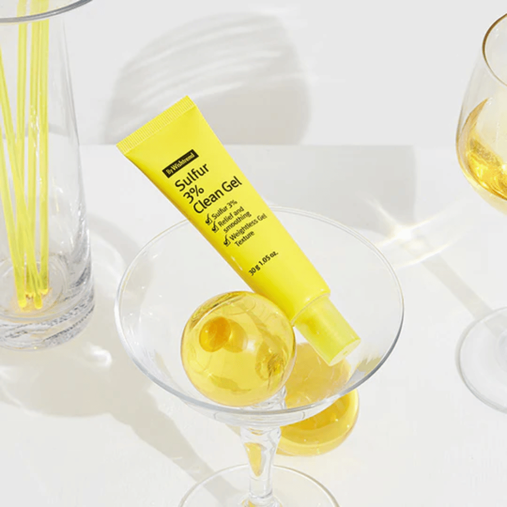 By Wishtrend Sulfur 3% Clean Gel keltaisessa tuubissa, joka on asetettu cocktail-lasiin keltaisen sfäärin kanssa, ympäröitynä tyylikkäillä laseilla valkoisella taustalla.