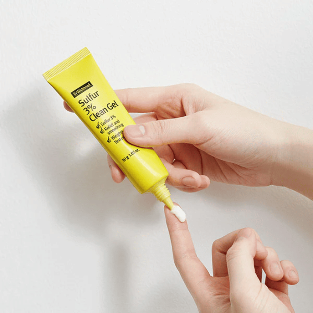 Lähikuva käsistä, jotka levittävät By Wishtrend Sulfur 3% Clean Gel, keltainen tuubi, jossa valkoinen geeli, valkoisella taustalla.