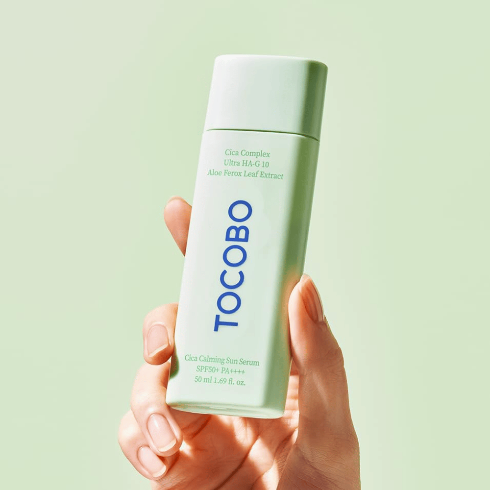 Käsi pitää TOCOBO Cica Calming Sun Serum -tuotetta mintunvihreässä pakkauksessa ja sinisellä tekstillä vaaleanvihreää taustaa vasten.