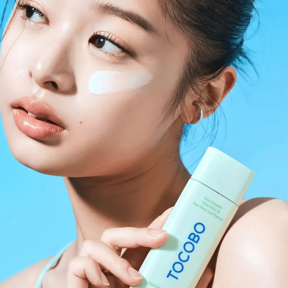Malli, jolla on aurinkovoidetta silmän alla, pitää TOCOBO Cica Calming Sun Serum -tuotetta mintunvihreässä pakkauksessa sinistä taustaa vasten.