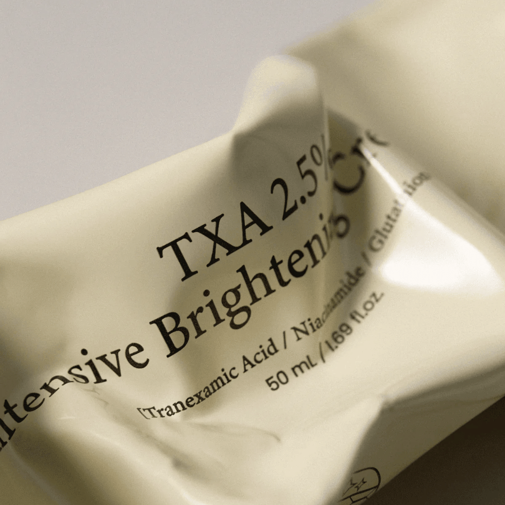 Lähikuva tuotteesta AXIS - Y TXA 2.5% Intensive Brightening Cream, jossa pehmeä ja minimalistinen tuubimuotoilu on keskiössä. Kuva korostaa tuotteen nimeä, Tranexamic Acidia ja Niacinamidea sekä eleganttia beigeä pakkausta.