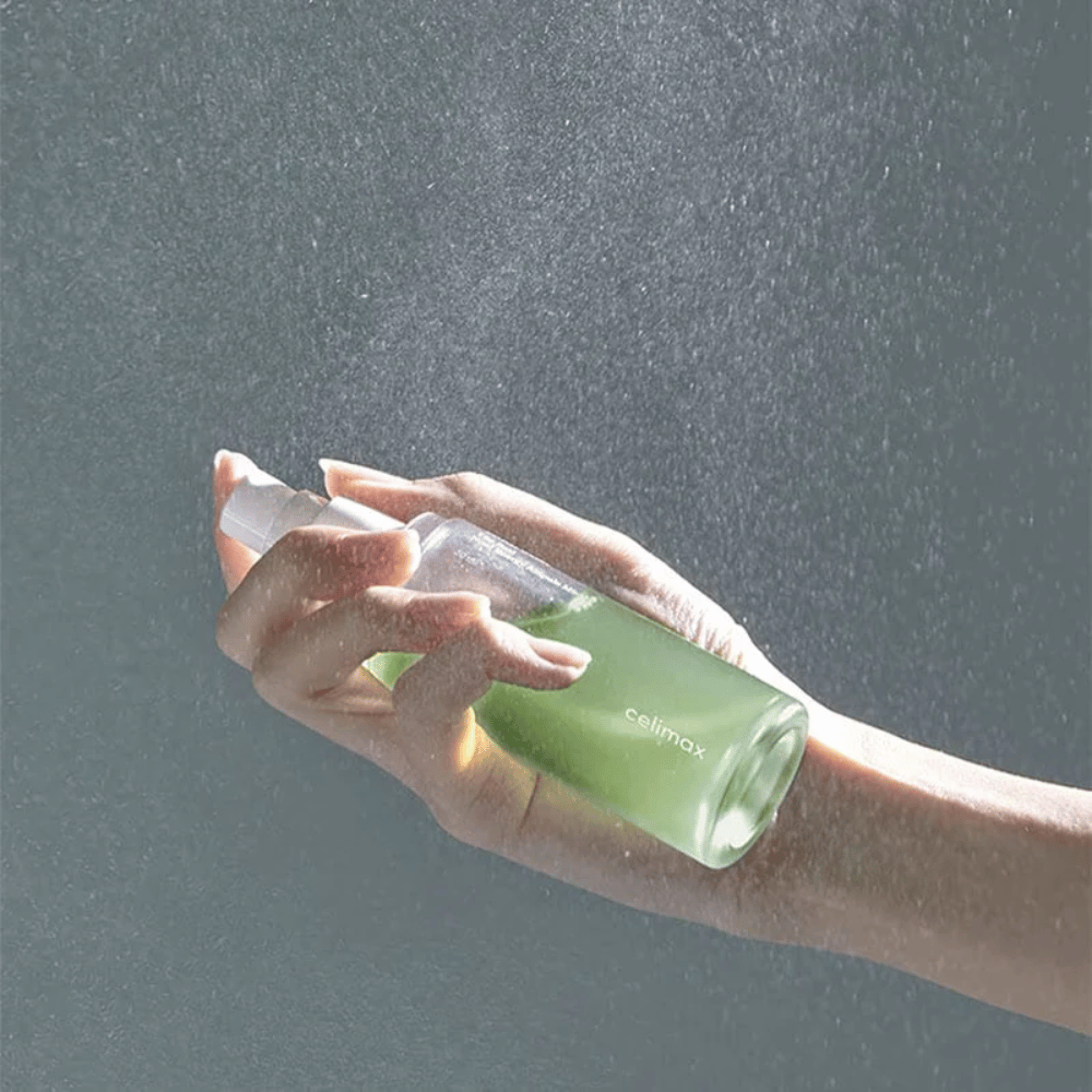 Käsi pitää Celimax Noni Mist -suihkepulloa ja levittää tuotetta hienolla, tasaisella suihkulla iholle. Pullo on vihreä ja tuotetta markkinoidaan nimellä "The Real Noni Energy Ampule Mist" antamaan energiaa ja kosteutusta iholle.