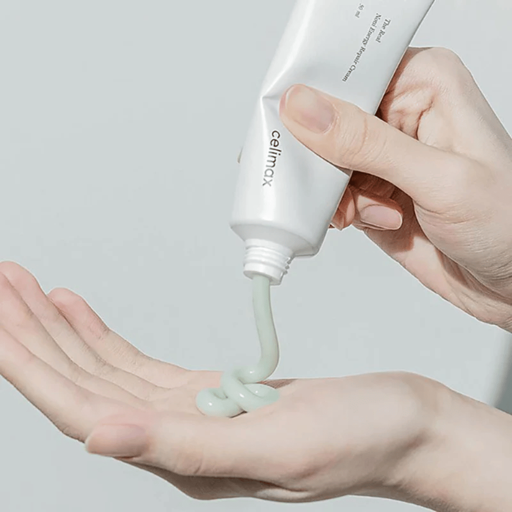 Käsi pitelee valkoista tuubia, jossa on Celimax Noni Energy Repair Cream, ja voidetta puristetaan tasaisena virtana kämmenelle. Voide on vaaleanvihreää ja markkinoidaan ravitsemaan ja palauttamaan ihoa.