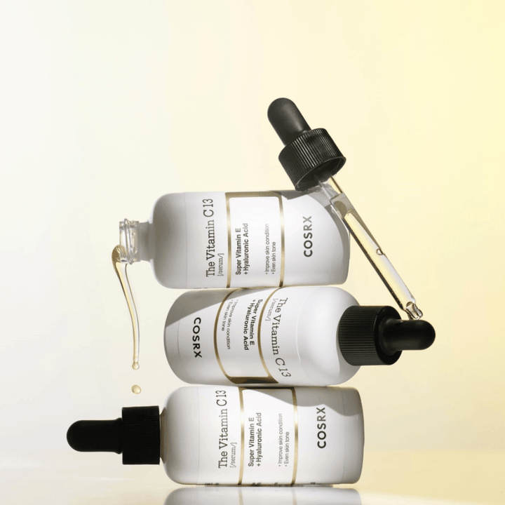 COSRX The Vitamin C 13 Serum E-vitamiinilla ja hyaluronihapolla, kolme pulloa pipetillä ja C-vitamiiniseerumin tippoja epätasaista ihonsävyä ja hehkua vastaan.