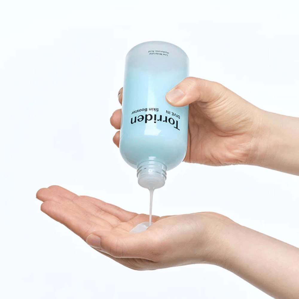 Kädet pitävät ihonhoitotuotepulloa ja annostelevat kirkasta nestettä kämmenelle. Pullossa on etiketti "Torriden - DIVE-IN Low Molecule Hyaluronic Acid Skin Booster".