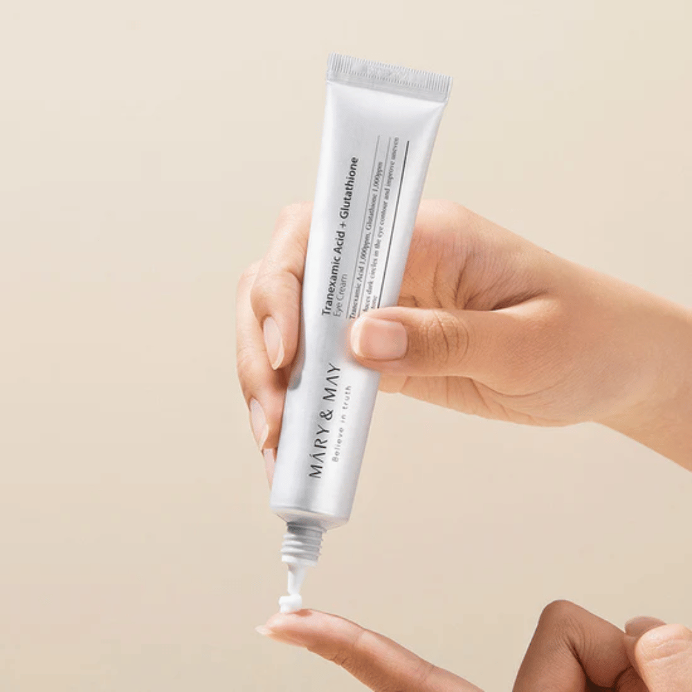 Käsi pitelee MARY & MAY -tuubia "Tranexamic Acid + Glutathione Eye Cream". Tuubi on valkoinen ja sen muotoilu on puhdas ja yksinkertainen, mikä korostaa tuotteen rauhoittavia ja kirkastavia ominaisuuksia silmänympärysalueelle. Tuote levitetään varovasti, keskittyen tarkkuuteen ja tehokkuuteen.