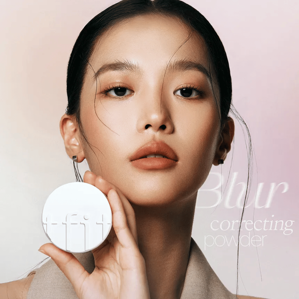 Nainen, jolla on virheetön meikki, pitää pyöreää valkoista rasiaa, jossa on TFIT-logo, ja taustalla teksti "Blur Correcting Powder".