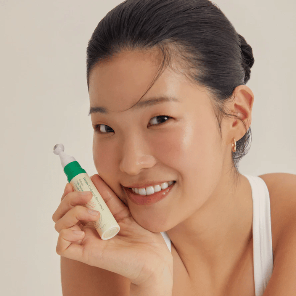 Nainen, jolla on ylös nostetut hiukset ja kultaiset korvakorut, pitää AXIS-Y Vegan Collagen Eye Serum 10ml lähellä kasvojaan, samalla kun hän hymyilee lempeästi. Seerumi on suunniteltu kosteuttamaan ja vähentämään ryppyjä silmien ympärillä, erityisesti herkälle iholle kehitetty.