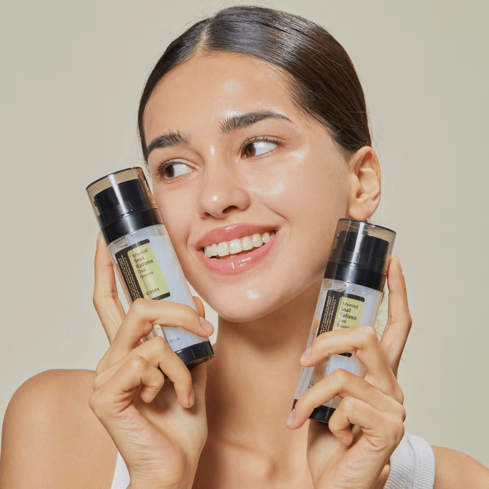 Hymyilevä nainen kiiltävällä iholla pitää kahta pulloa COSRX Advanced Snail Radiance Dual Essence 80 ml lähellä kasvojaan vaaleaa taustaa vasten.