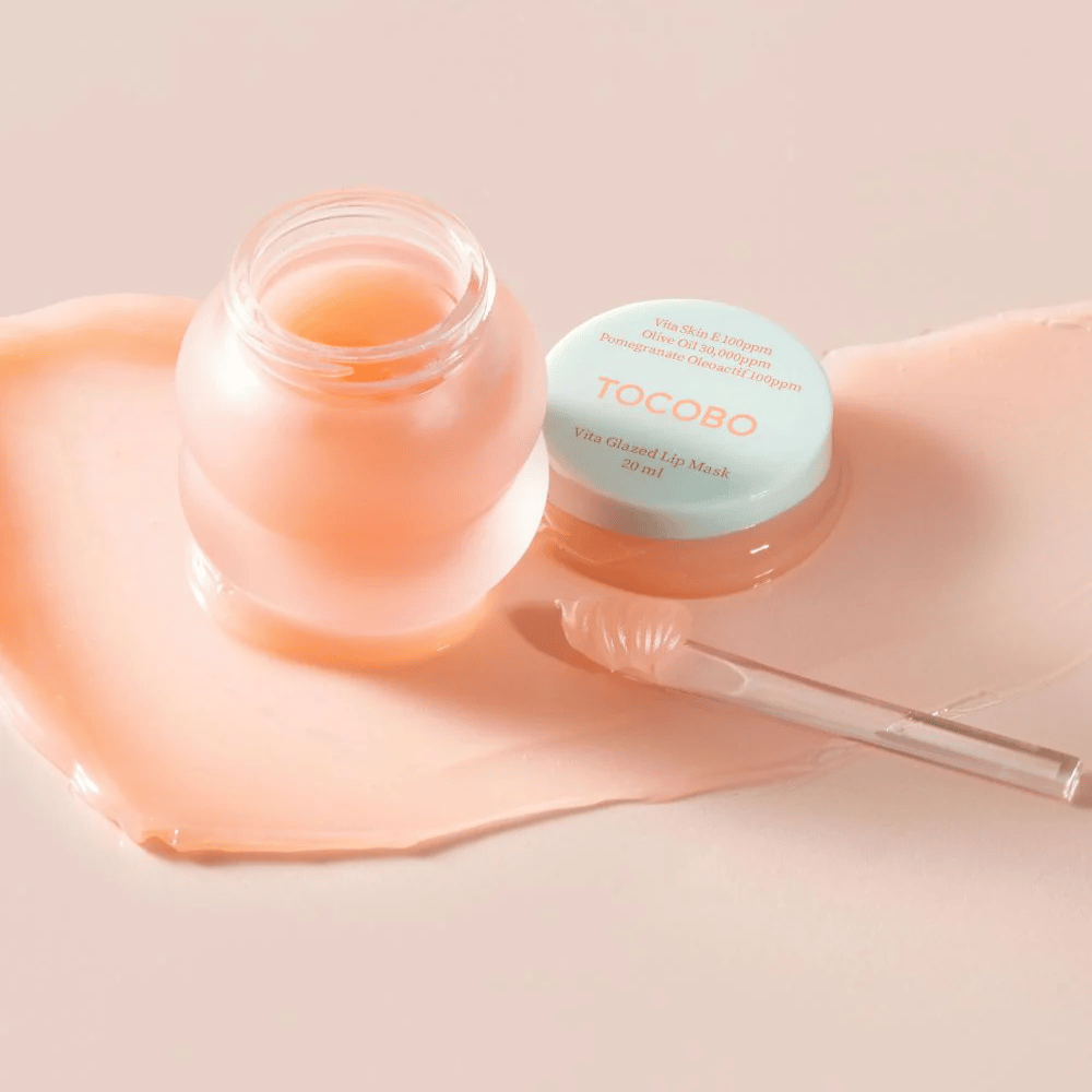 Näkyvissä on purkki, jossa lukee "Vita Glazed Lip Mask" Tocobo. Purkki on läpinäkyvä, vaalean turkoosilla kannella ja sisältää kiiltävän, vaaleanpunaisen huulinaamion. Pienen lastan vieressä on purkki, ja osa naamiosta on levitetty alustalle. Tuote on suunniteltu kosteuttamaan ja hoitamaan huulia.