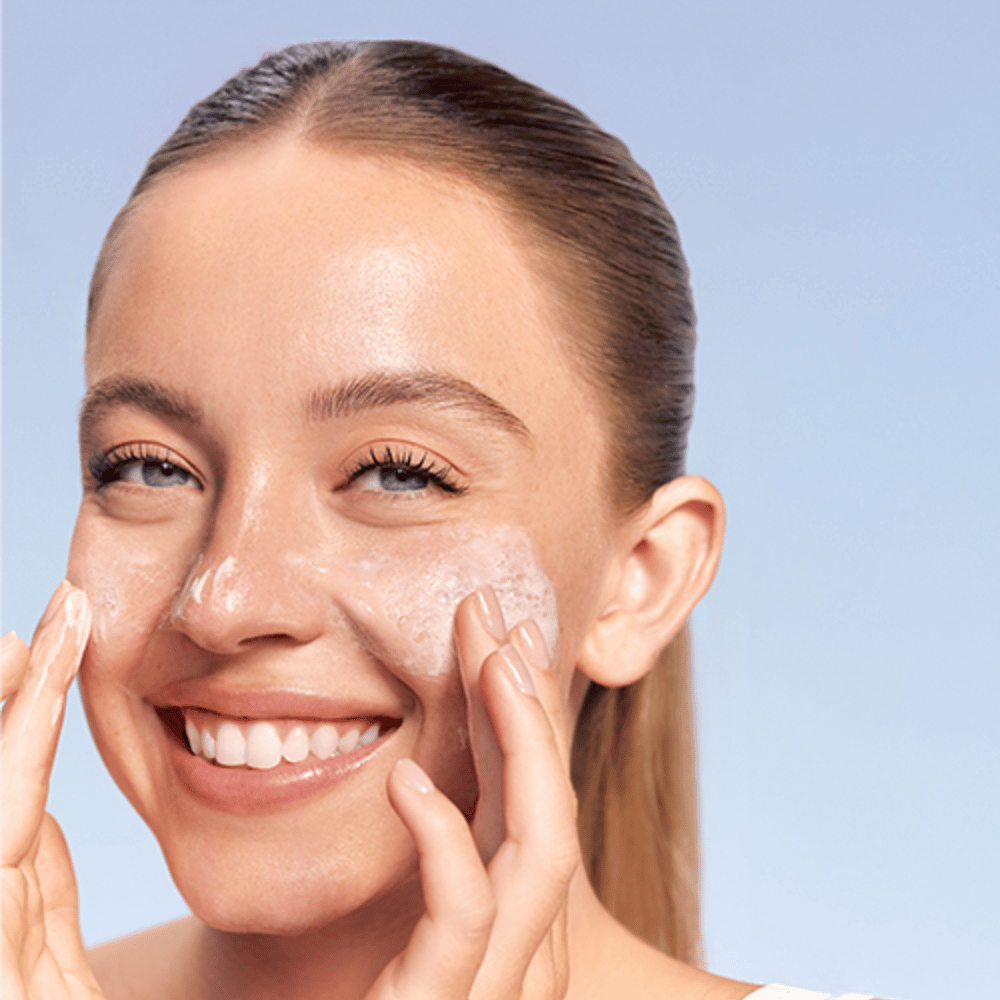 Hymyilevä nainen levittää LANEIGE Water Bank Gentle Gel Cleanser vaahtoavana poskilleen, sinistä taustaa vasten.