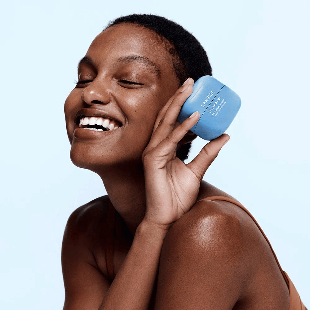 Hymyilevä nainen pitää sinistä purkkia LANEIGE Water Bank Blue Hyaluronic Cream poskeaan vasten, vaaleansinistä taustaa vasten.