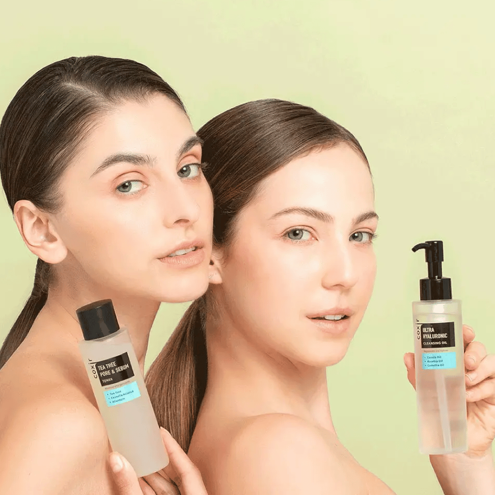 Kaksi naista, joilla on paljaat hartiat, poseeraa ihonhoitotuotteiden kanssa; toinen pitää kädessään pulloa, jossa on merkintä "TEA TREE PORE & SEBUM", ja toinen pumppupulloa, jossa on merkintä "ULTRA HYALURONIC CLEANSING OIL". He seisovat vaaleanvihreän taustan edessä.