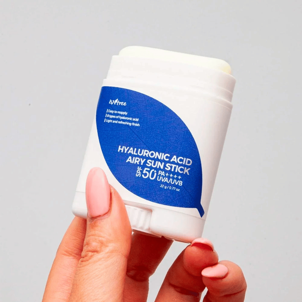 Käsi pitää valkoista aurinkopuikkoa, jossa on sininen etiketti merkiltä Isntree. Tuotteen nimi on "Hyaluronic Acid Airy Sun Stick" ja siinä on aurinkosuojakerroin SPF 50+ PA++++.