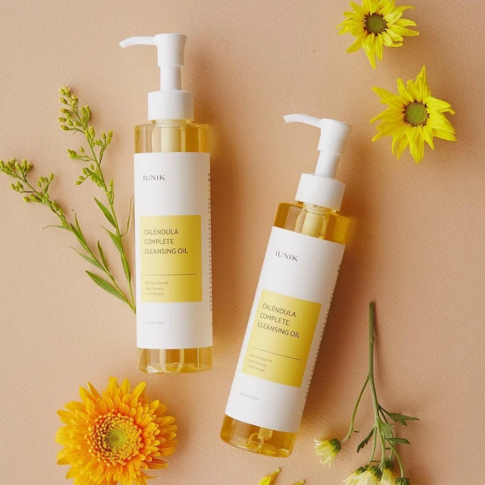 Kaksi pulloa iUNIK Calendula Complete Cleansing Oil 200ml, joissa on keltaisia kukkia beigellä taustalla, infusoitu kasviöljyllä.