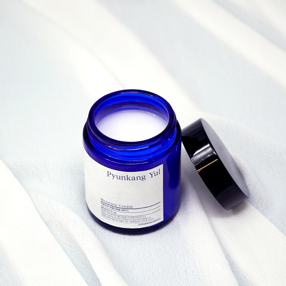Sininen purkki kannella valkoisen liinan päällä esittelee Pyunkang Yul'n Moisture Cream 100 ml kosteuttavia ominaisuuksia.
